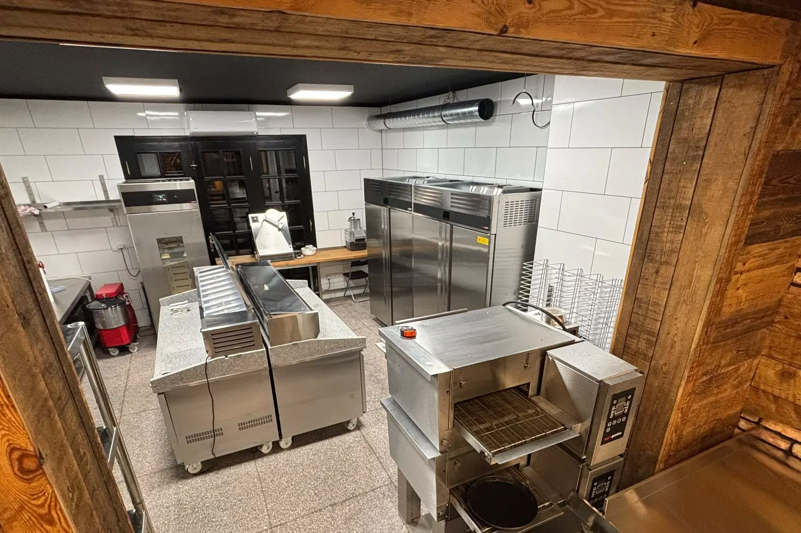 Kuchnia pizzerii z piecem tunelowym, widziana przez drewniane przejście